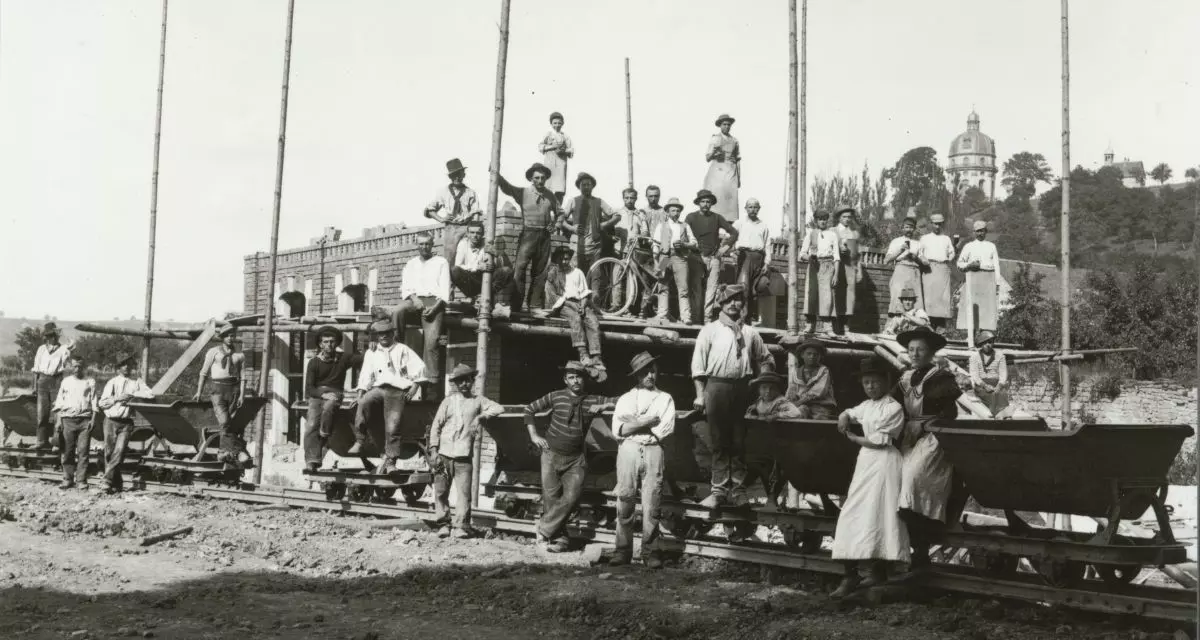Schwarz-weiße Fotografie einer großen Gruppe von Männern, Frauen und Kindern auf einem Baugerüst und Loren. Die Personen tragen Arbeitskleidung aus dem frühen 20. Jahrhundert. Im Hintergrund ist ein im Bau befindliches Backsteingebäude zu sehen, weiter hinten eine Kirche mit Kuppel auf einem Hügel.