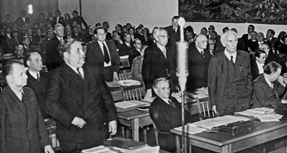 Carlo Schmid (2. v. l. stehend, vorne) im Parlamentarischen Rat in Bonn, 1949. Rechts im Bild auch Theodor Heuss, ein weiterer maßgeblicher Vater des Grundgesetzes. (Foto: LMZ BW) Schwarz/Weiß-Foto von Raum mit Tischen und Stühlen. An denen sitzen bzw stehen Männer in Anzügen.