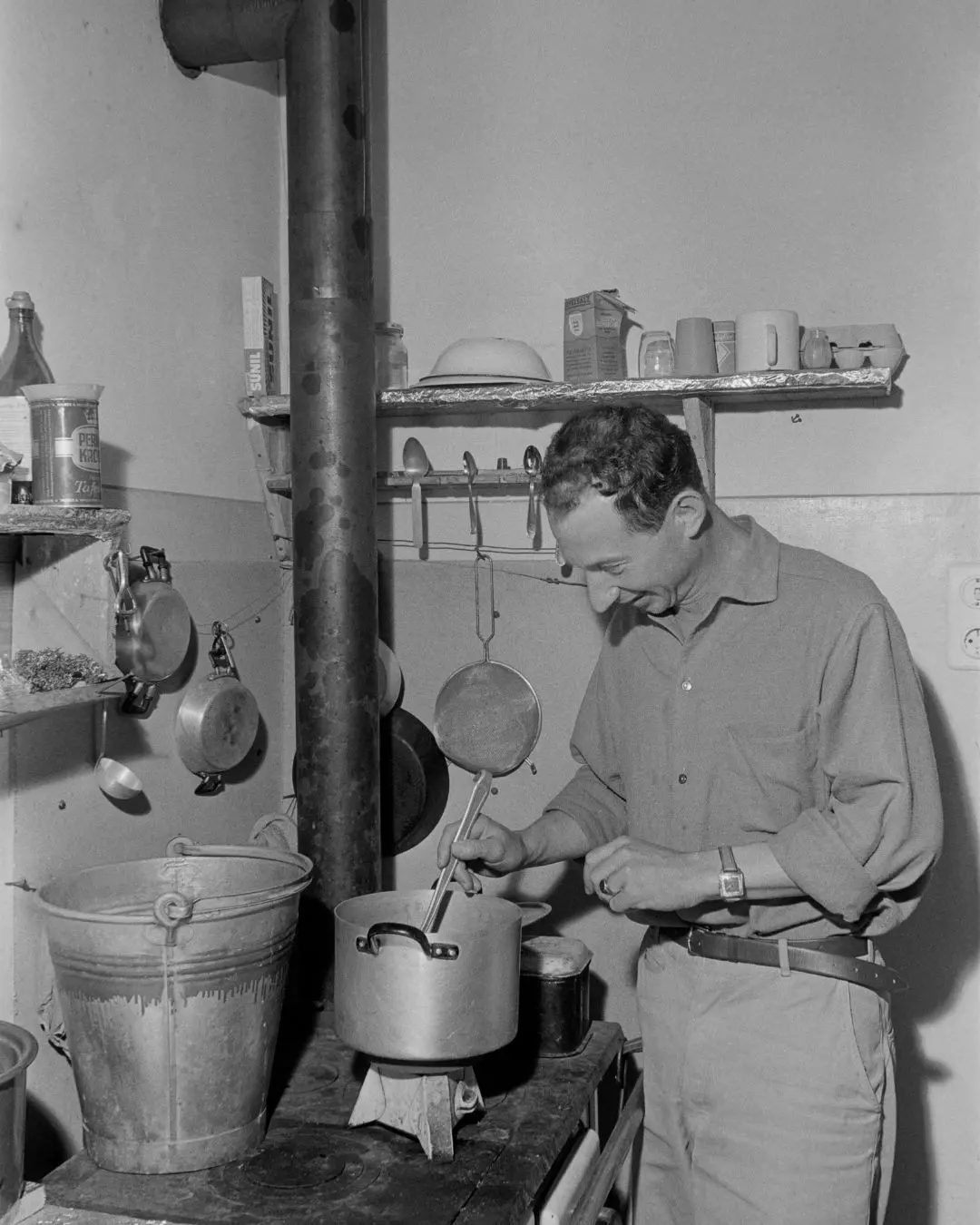 Schwarz-weiß Foto von Mann am alten Herd mit großen, alten Töpfen. Mann rührt im hinteren Topf.