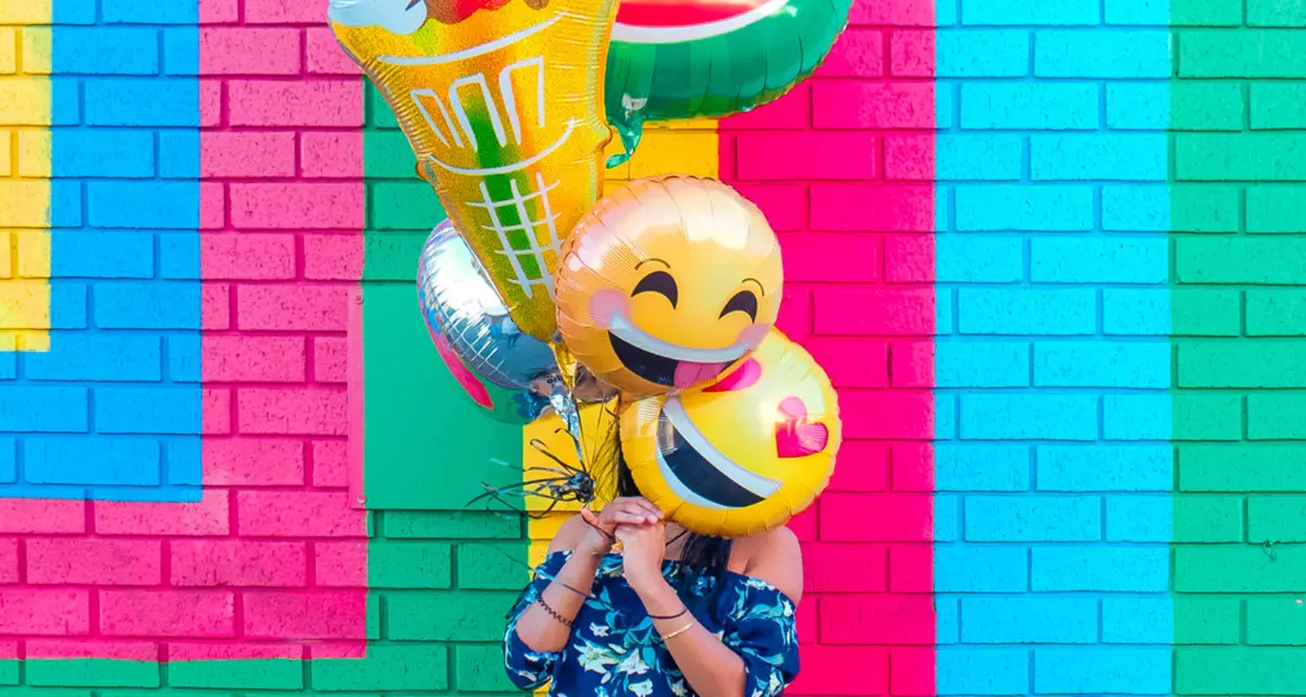 Mädchen vor farbiger Hauswand mit bunten Luftballons vor dem Gesicht