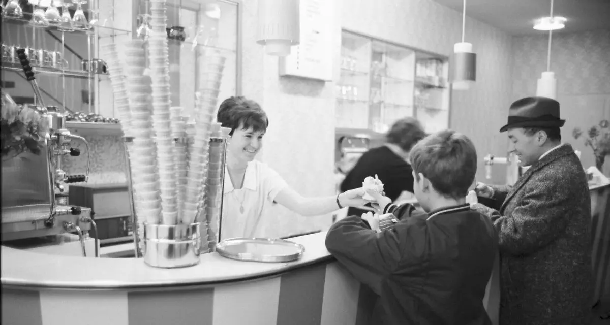 Schwarz-weiß Foto von einer Eisdiele. Hinter der gestreiften Theke steht eine Frau, die einem Kind vor der Theke ein Eis im Pappbecher gibt.