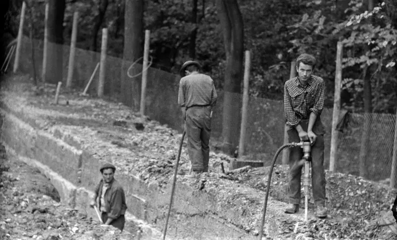 Schwarz-weiß Foto von Baustelle mit Mauer und Zaun. Drei Männer arbeiten dort ohne Schutzkleidung und mit schwerem Gerät.