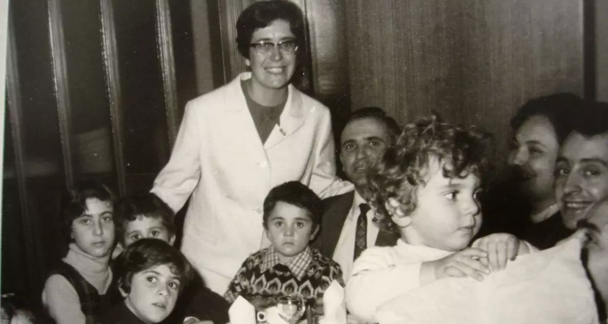 Schwarz-weiß Foto von einer Frau umgeben von Kindern und einigen Erwachsenen im Hintergrund. Die Frau strahlt in die Kamera.