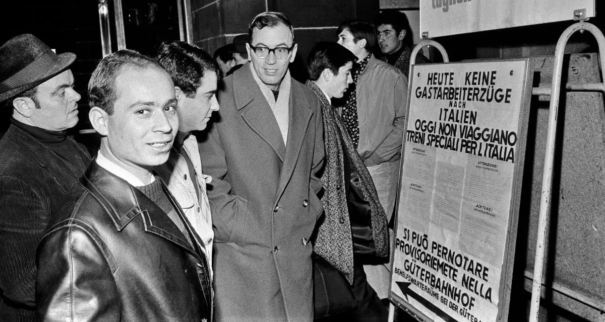 Schwarz-weiß Foto von Männern die vor einer Tafel stehen auf der deutscher und italienischer Text steht. Der Mann vorne im Bild schaut offen in die Kamera, die Männer scheinen sich zu unterhalten.