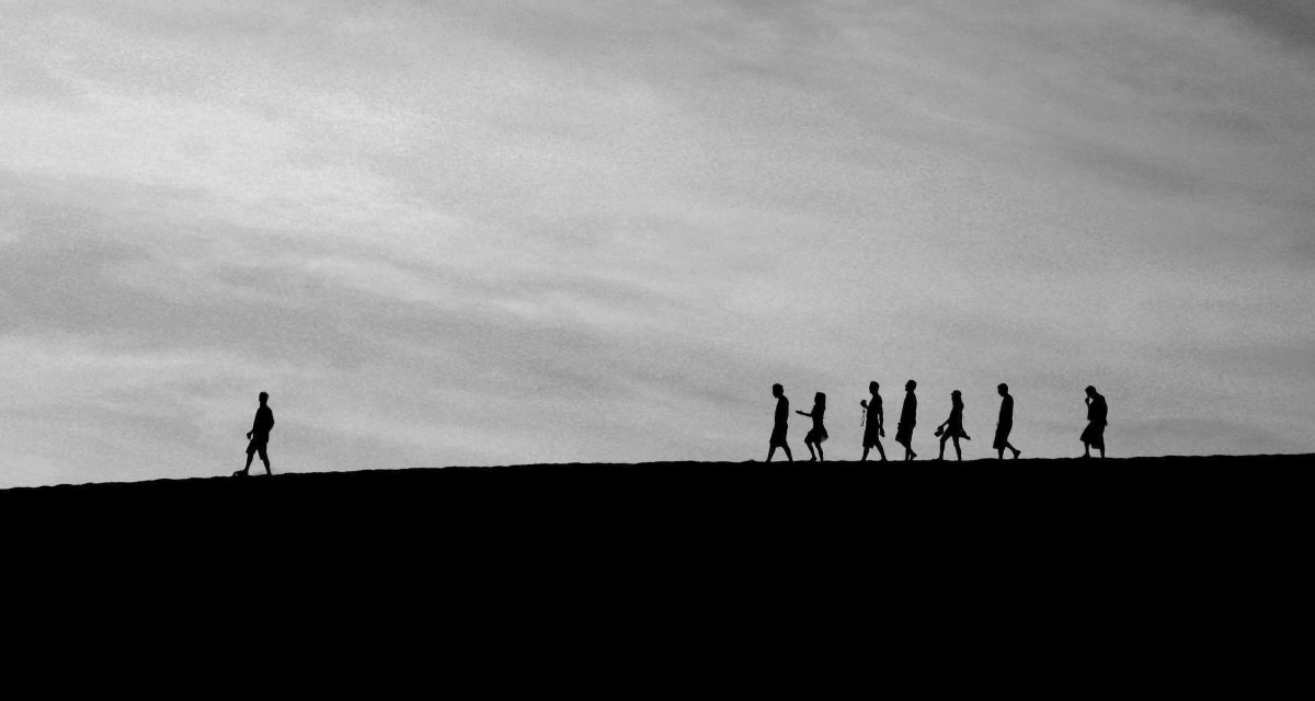 Silhouetten von Menschen. Ein Mensch geht in eine Richtung, viele weitere gehen als Gruppe dahinter.