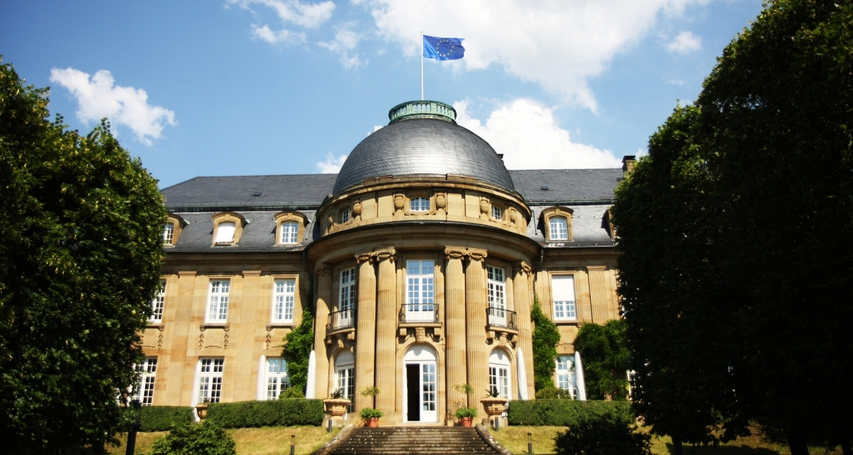 Villa Reitzenstein in Frontalansicht und Sonnenbeschienen. Sandsteinfarbenes Gebäude mit grauem Dach. Vorne in der Mitte runde Kuppel mit EU-Flagge.