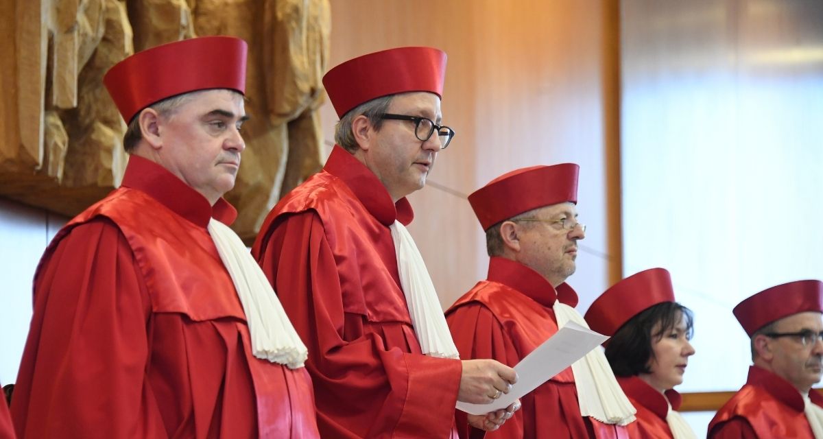 Foto von Richter:innen in roten Roben und Hüten vor einer Holzwand. Einer hält ein Papier in der Hand.