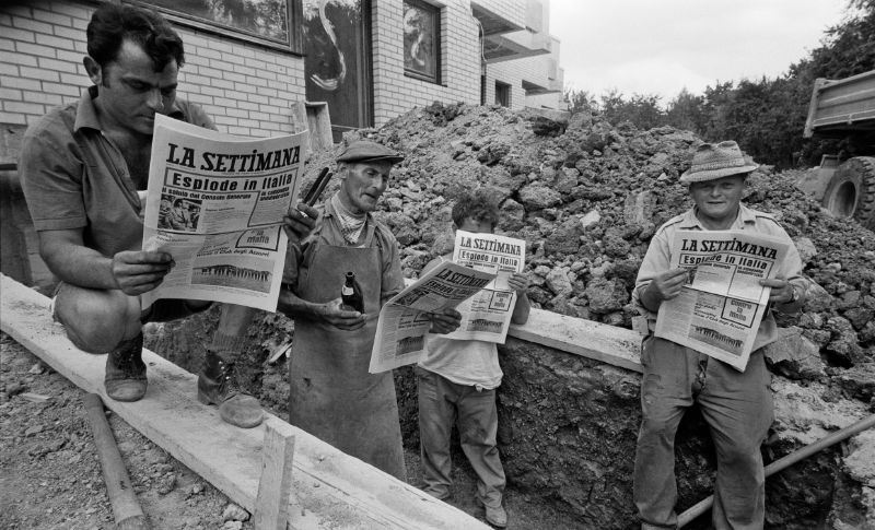 Schwarz-weiß Foto von vier Männern, die auf einer Baustelle stehen. Alle halten eine Ausgabe der gleichen italienischen Zeitung in der Hand und lesen sie. 